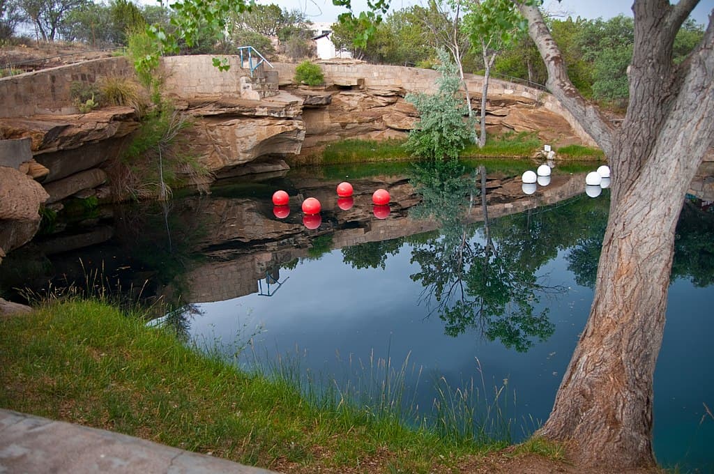 View of the Blue Hole in Santa Rosa, NM near Route 66 with red and white dive buoys on the calm surface and a tree in the foreground to indicate one of the most enjoyable backcountry swimming holes in the country