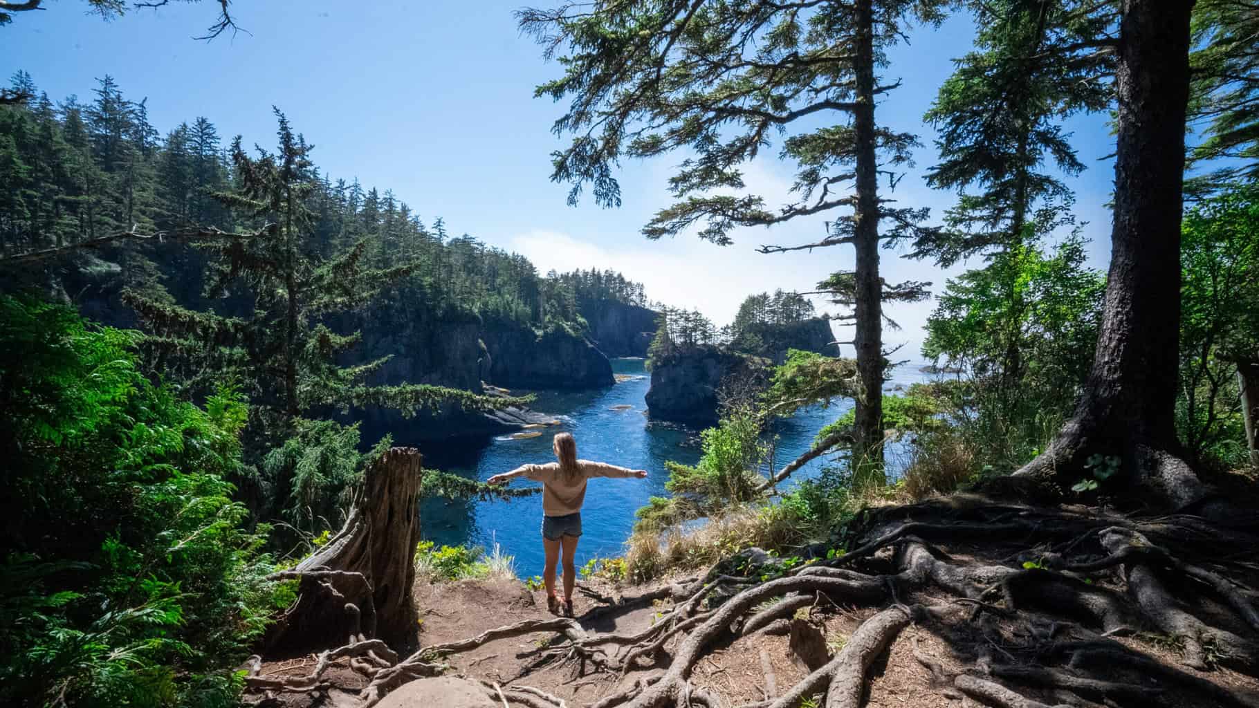Alice Ford with arms outstretched overlooking Cape Flattery through evergreen trees during a 4-day hiking trip solo through Olympic National Park
