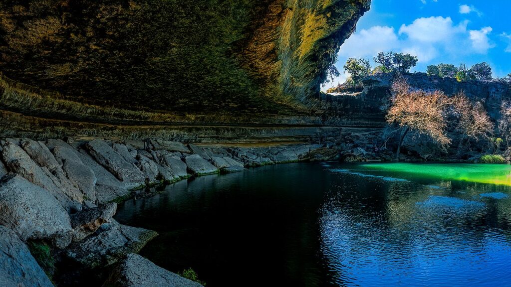 View of Hamilton Pool in Dripping Springs, Texas on a sunny day with the rock ledge creating shade and brightly-colored water in the swimming hole