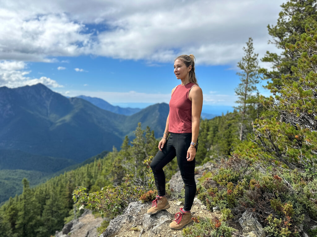 Alice Ford standing exhilarated at the top of Mt. Townsend with clouds overhead, coniferous trees around her, and mountain peaks from the Olympic range in the distance after a hike to the summit as part of a 4-day itinerary in Olympic National Park