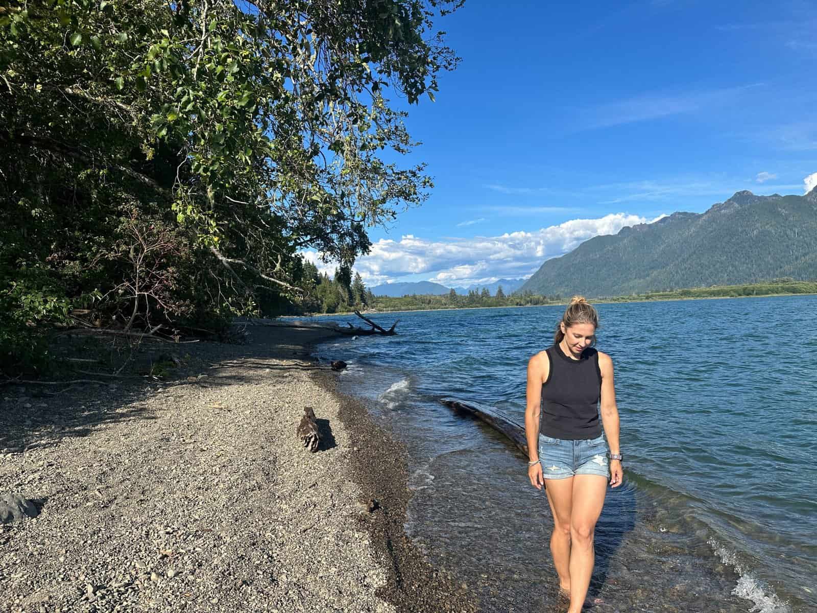 Alice Ford strolls along the shore of Lake Quinault during the final day of her 4-day itinerary for Olympic National Park with deciduous trees around her and mountain views across the water