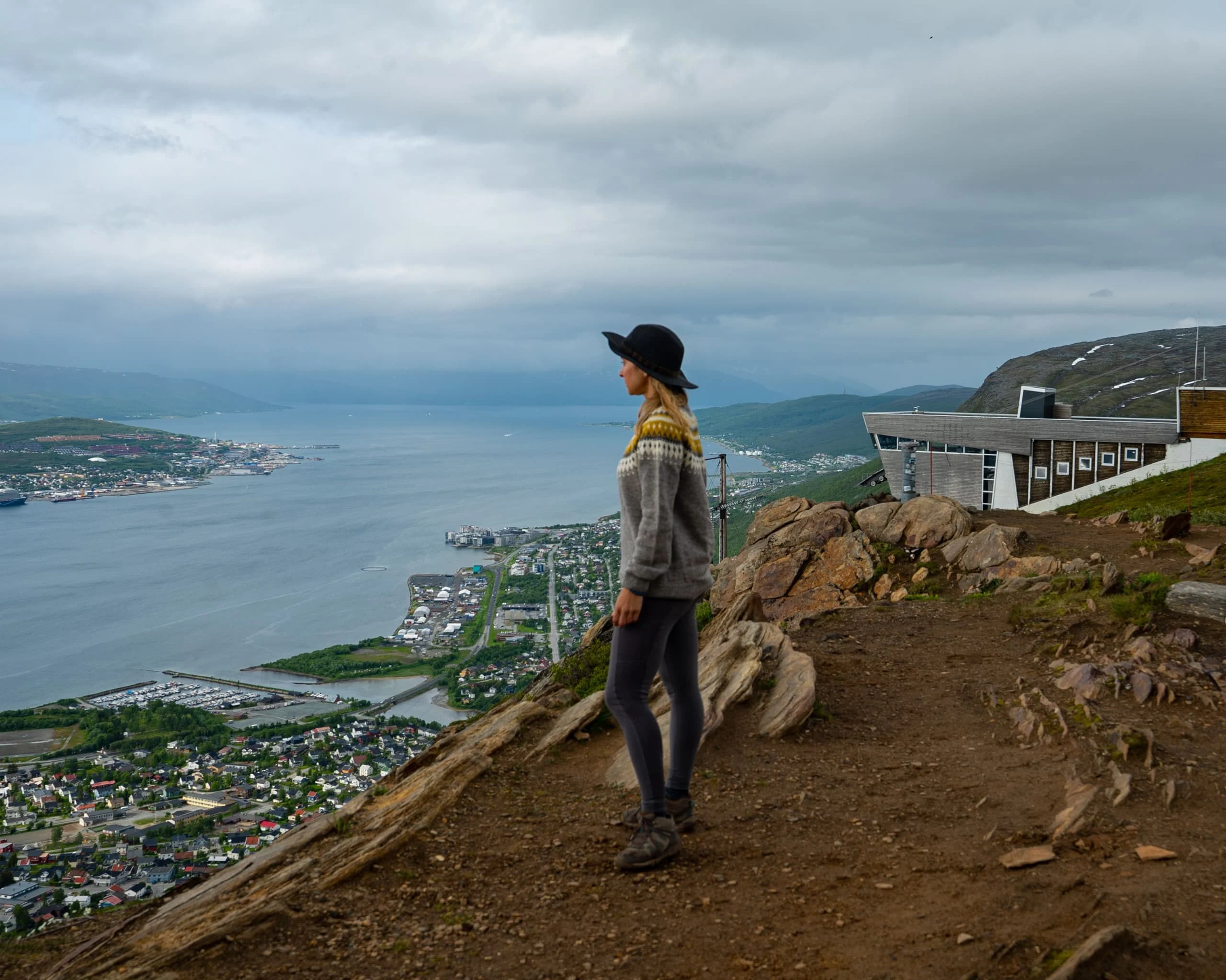 Alice standing on a cliff edge above a fjord and a distant harbour