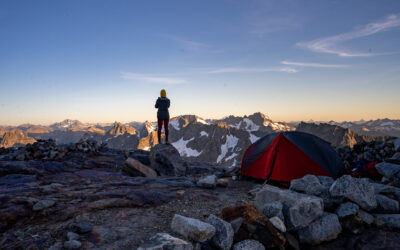 Woman stands on a rocky outcropping on top of a mountain next to a tent as the sun sets