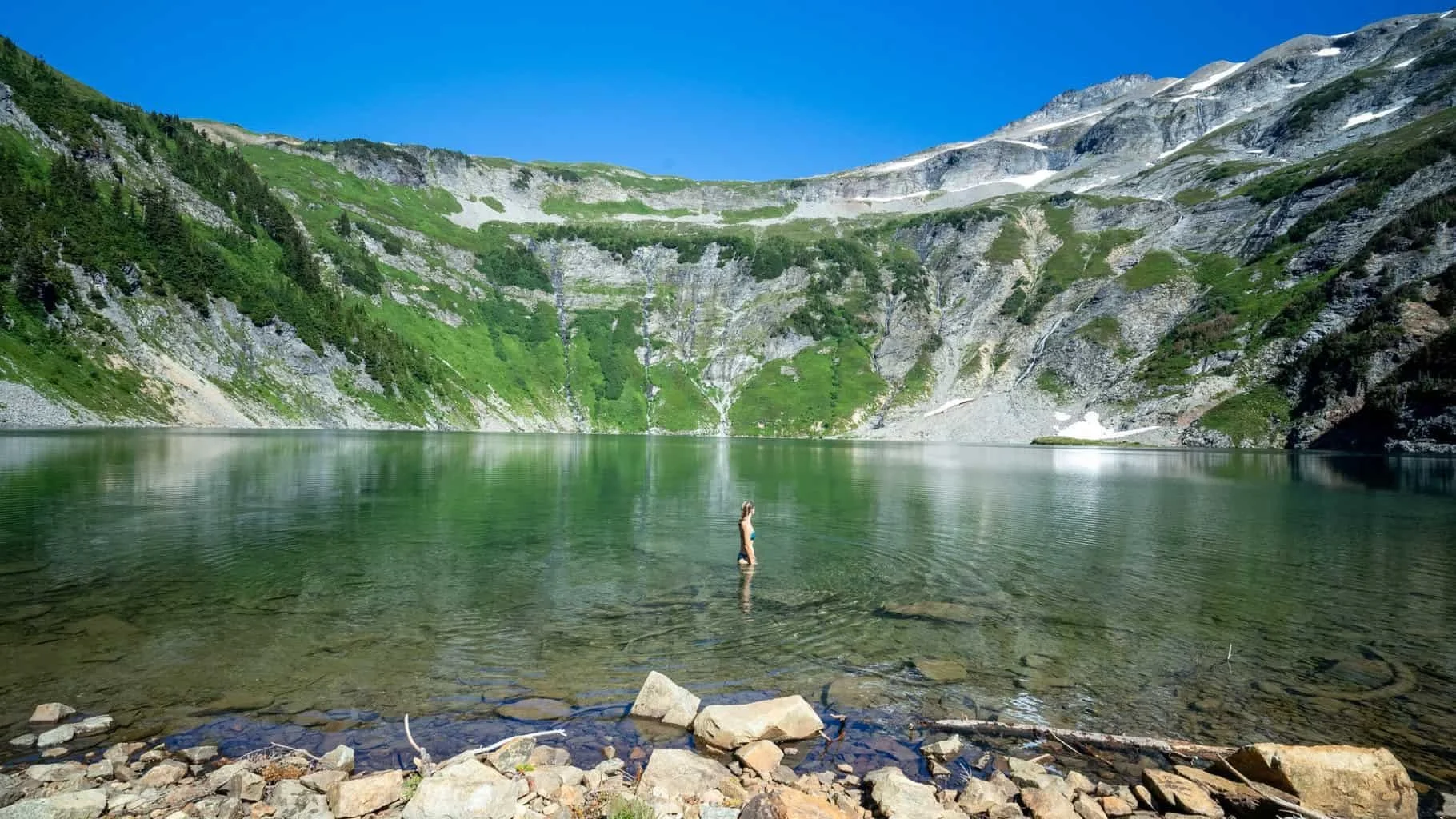Alice wading into a turquoise glacial lake beneath sheer, forested cliffs in the North Cascades