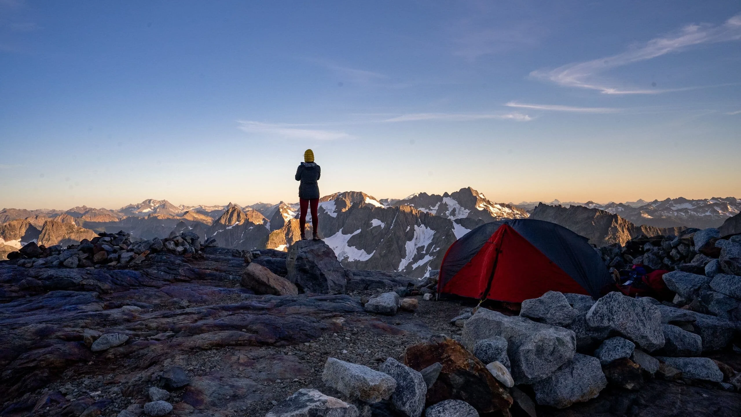 Dawn in the North Cascades: Alice stands on a granite ridge beside a red tent, glaciated peaks fading into the distance