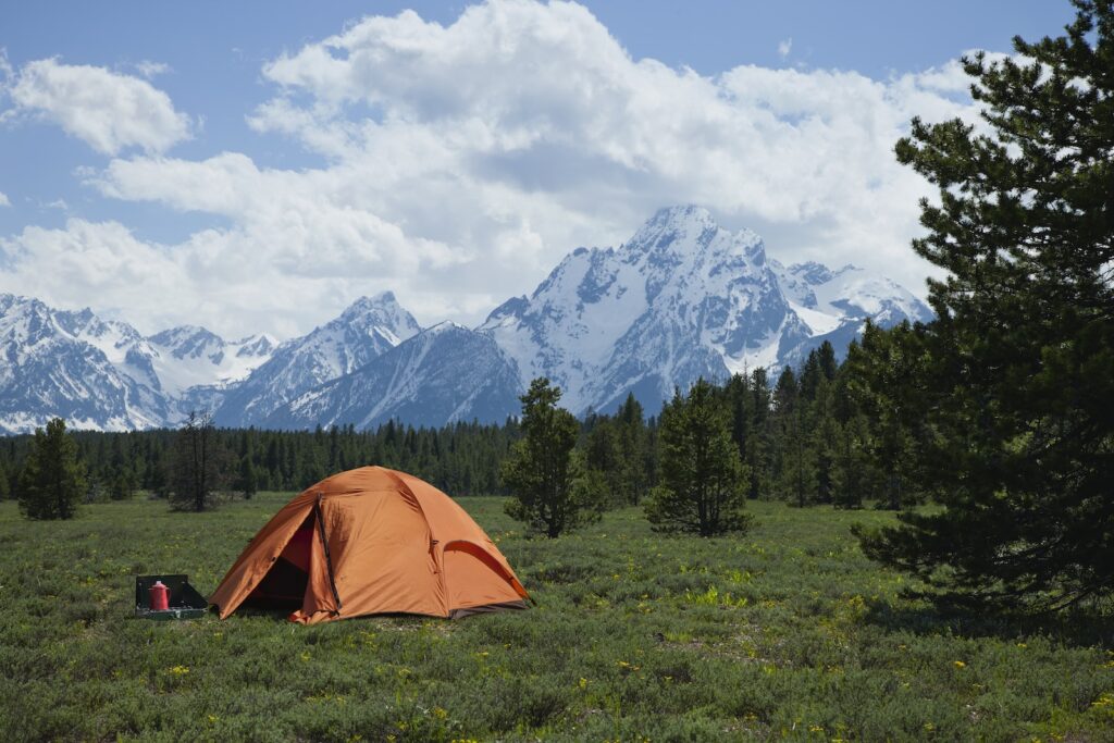 Orange tent set up in a meadow in the shadow of Grand Teton Mountains to demonstrate how to plan lodging or camping accommodations on a U.S. national park road trip