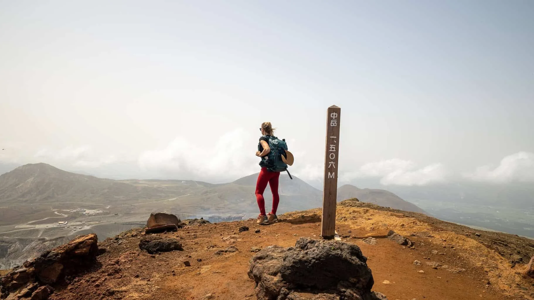 A hiker at a summit marker on a volcanic ridge in Kyushu, Japan