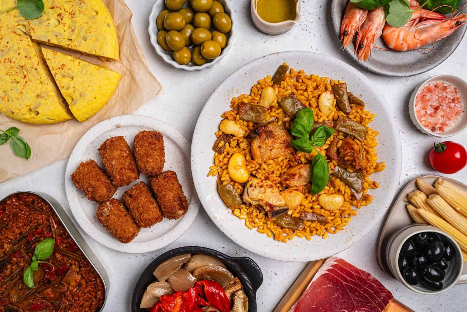 Spanish food table paella valenciana, croquettes, omelette, and tapas slices jamon, bowls with olives, spicy potatoes, shrimp. Typical spanish dishes, tapas set concept. Variety of plates